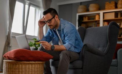Man sitting at laptop with credit card in hand looking worried. Adobe.